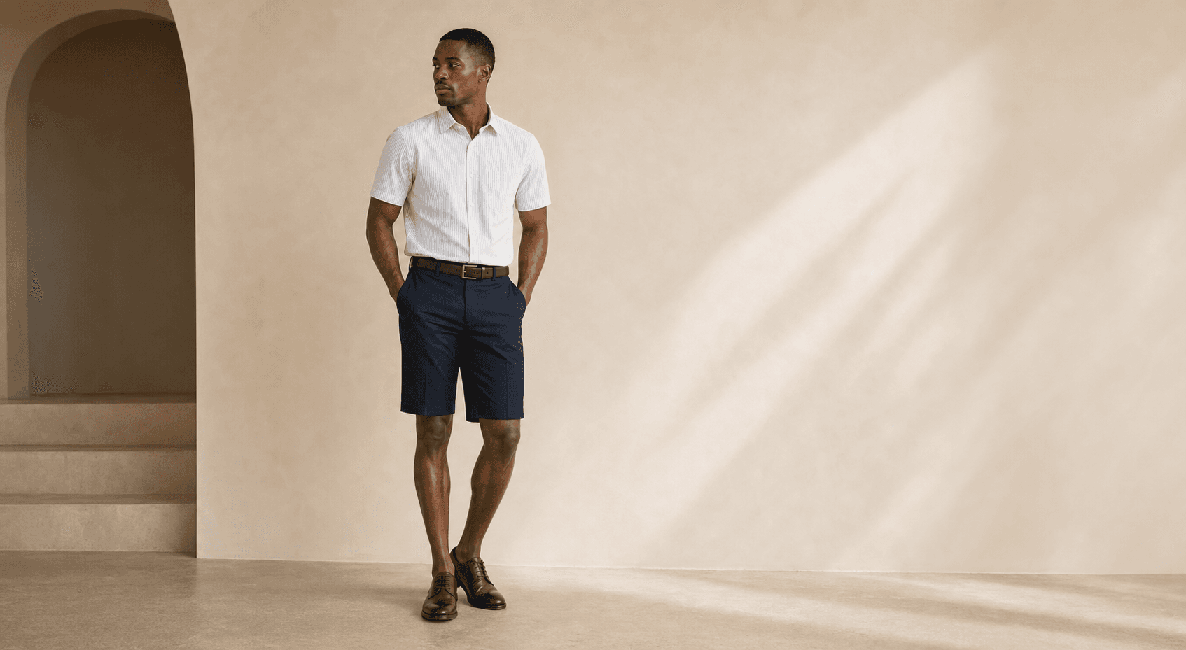 Black male model wearing a short sleeve shirt, tailored shorts, belt, and dress shoes